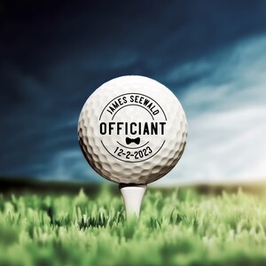May include: A white golf ball with a black circle on it that says "James Seewald Officiant 12-2-2023" with a small bow tie graphic.