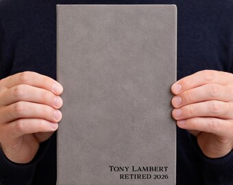 May include: A gray, rectangular journal with a textured cover is held by a person. The journal has the text "TONY LAMBERT RETIRED 2026" printed in black at the bottom. The person is wearing a dark blue sweater.