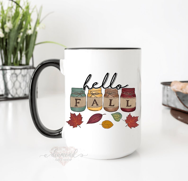May include: White ceramic mug with a black handle. The mug has a design of four mason jars with the word "FALL" spelled out in the jars. The jars are decorated with burlap and twine. The text "hello" is written above the jars. There are also fall leaves around the jars.