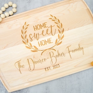 May include: A wooden cutting board with a decorative engraved design that says "Home Sweet Home" with a laurel wreath around the text. The board also has the text "The Diwarco-Baker Family Est. 2023" engraved on it.
