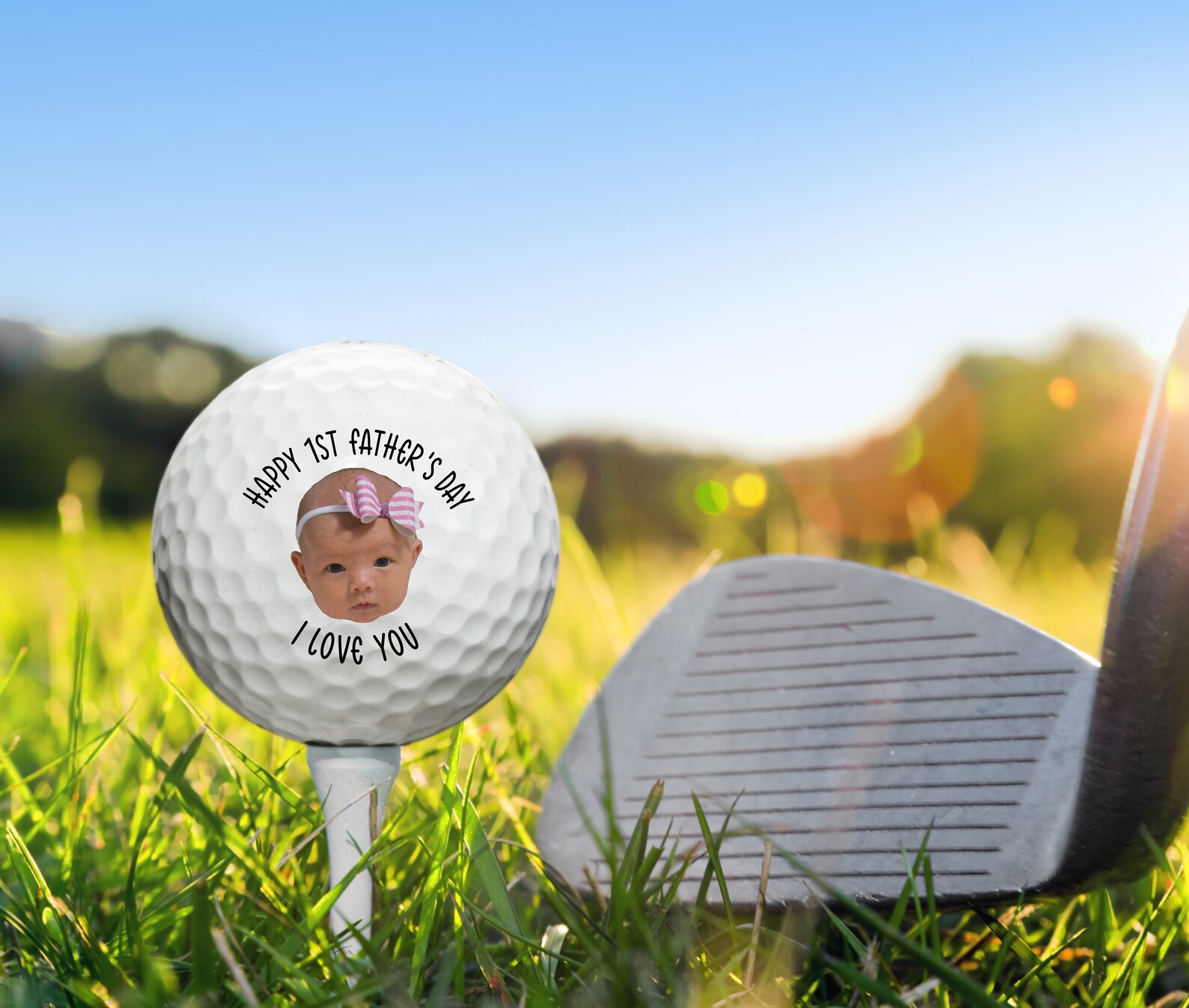 May include: A white golf ball with a baby's face and a pink bow on top. The text on the golf ball reads 'Happy 1st Father's Day I Love You'.