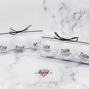 Pode incluir: Duas caixas de pl&aacute;stico transparente com la&ccedil;os de fita preta, cada uma contendo tr&ecirc;s bolas de golfe brancas com texto preto. O texto nas bolas de golfe diz "Ryan, Groomman" e "Carm, Groomman 11.3.2022".