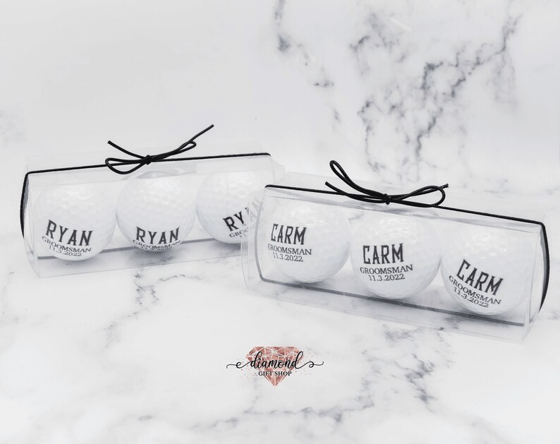 Pode incluir: Duas caixas de pl&aacute;stico transparente com la&ccedil;os de fita preta, cada uma contendo tr&ecirc;s bolas de golfe brancas com texto preto. O texto nas bolas de golfe diz "Ryan, Groomman" e "Carm, Groomman 11.3.2022".