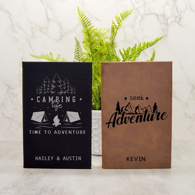 May include: Two personalized leather journals with a camping theme. One journal is black with silver text and a camping scene with the text "*CAMPING* life TIME TO ADVENTURE" and the names "HAILEY & AUSTIN". The other journal is brown with black text and a mountain scene with the text "*seek* Adventure" and the name "KEVIN".