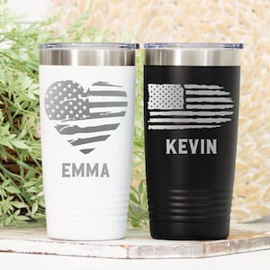 May include: Two stainless steel tumblers with a distressed American flag design. One tumbler is white with a heart-shaped flag and the name "EMMA" engraved on it. The other tumbler is black with a rectangular flag and the name "KEVIN" engraved on it.
