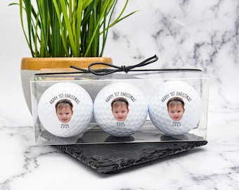 May include: Three white golf balls in a clear box, each with a baby's face and the text "HAPPY 1ST CHRISTMAS 2025". The box is tied with a black ribbon. A small potted plant is in the background.