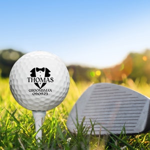 K&ouml;nnte beinhalten: Ein wei&szlig;er Golfball mit einer schwarzen Smoking-Grafik und dem Text "THOMAS GROOMSMAN 09.09.23".