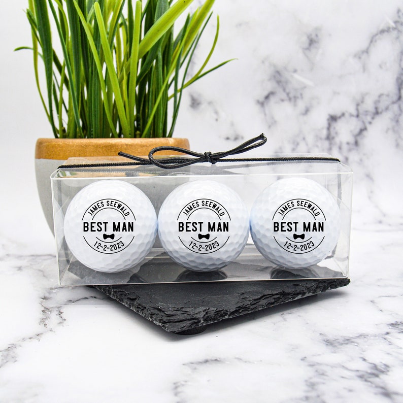 May include: Three white golf balls with black text and a bowtie design. The text reads "JAMES SEEWALD BEST MAN 12-2-2023". The golf balls are in a clear plastic box with a black ribbon.