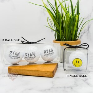 Pode incluir: Tr&ecirc;s bolas de golfe brancas com texto preto em uma embalagem de pl&aacute;stico transparente. O texto nas bolas de golfe diz "Ryan, Groomman, 11.3.2022". Uma &uacute;nica bola de golfe branca com um rosto sorridente amarelo em uma embalagem de pl&aacute;stico transparente. O texto "3 Ball Set" e "Single Ball" est&aacute; impresso abaixo da embalagem.