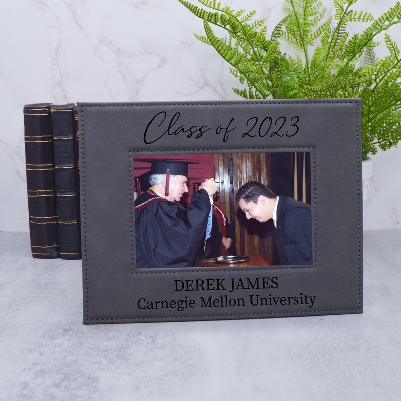 May include: Gray leather photo frame with a stitched border. The frame has a black inscription that reads "Class of 2023". A photo of a graduation ceremony is in the frame. The inscription below the photo reads "Derek James Carnegie Mellon University".