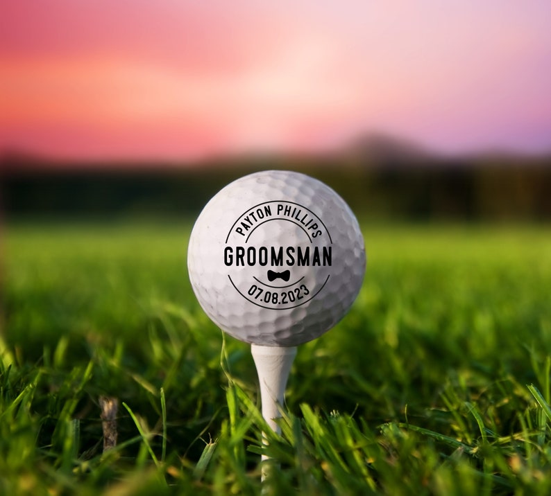May include: A white golf ball with a black circle sticker that says "PAYTON PHILLIPS GROOMSMAN 07.08.2023" with a bow tie icon.