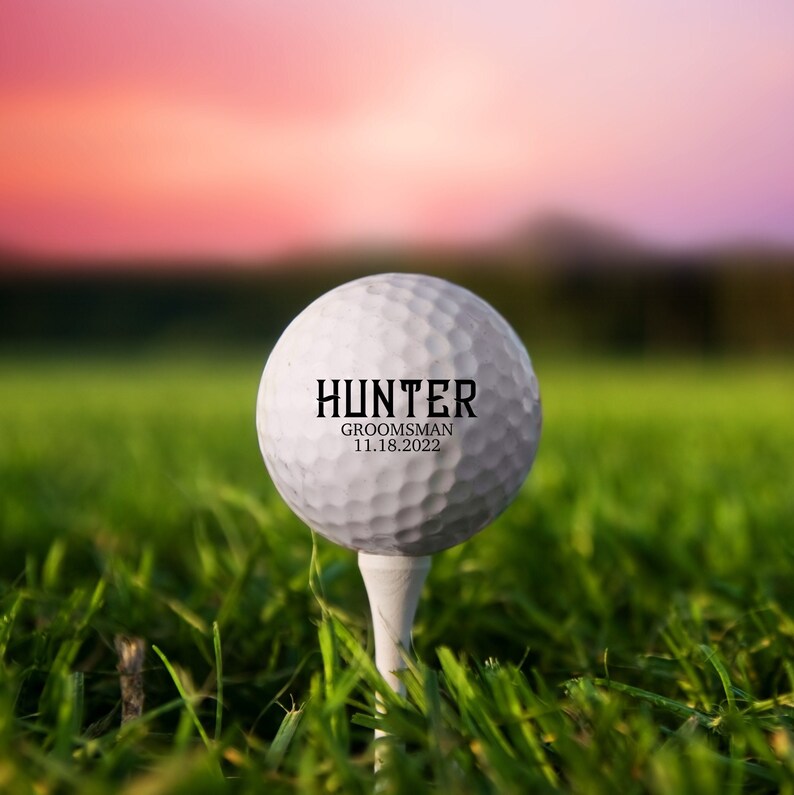 Pode incluir: Uma bola de golfe branca com o texto "HUNTER GROOMSMAN 11.18.2022" impresso nela, sentada em um tee de golfe verde em um campo gramado.