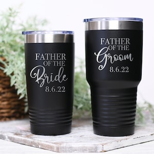 May include: Two black stainless steel tumblers with a silver inscription. The taller tumbler says "Father of the Groom 8.6.22" and the shorter tumbler says "Father of the Bride 8.6.22".