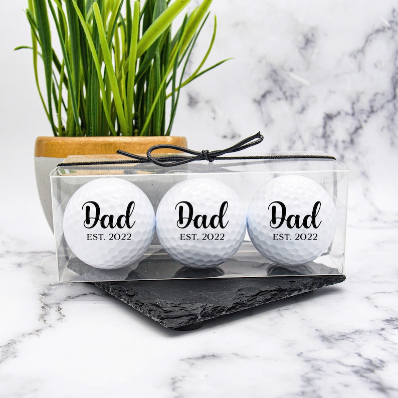 May include: Three white golf balls in a clear plastic box with a black ribbon. Each ball has the word "Dad" printed on it with the year "EST. 2022" underneath.
