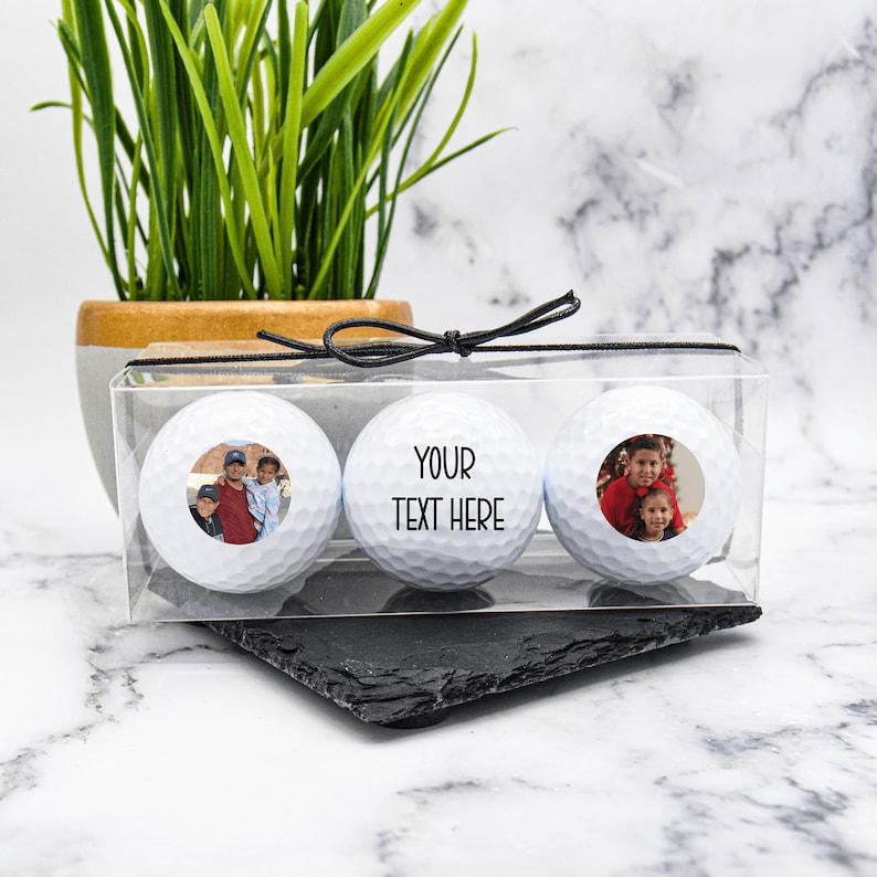 May include: Three white golf balls in a clear plastic box with a black ribbon. Each ball has a photo of a family on it. The middle ball has the text "YOUR TEXT HERE" on it.