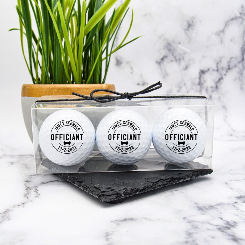 May include: Three white golf balls in a clear plastic box with a black ribbon. Each golf ball has a black and white sticker with the text "James Seewald Officiant 12-2-2023" and a small bow tie graphic.