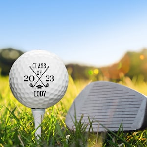 May include: A white golf ball with black text that reads "Class of 2023 Cody" with two crossed golf clubs and a graduation cap. The golf ball is on a tee in green grass with a golf club in the background.