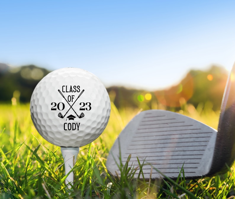 May include: A white golf ball with black text that reads "Class of 2023 Cody" with two crossed golf clubs and a graduation cap. The golf ball is on a tee in green grass with a golf club in the background.