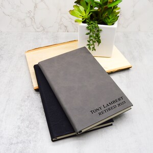 May include: Two leather-bound journals, one grey and one black, with the text "TONY LAMBERT RETIRED 2023" embossed on the grey journal.