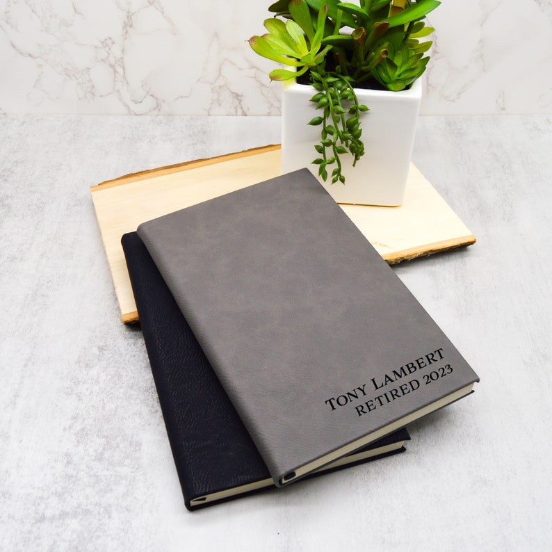 May include: Two leather-bound journals, one grey and one black, with the text "TONY LAMBERT RETIRED 2023" embossed on the grey journal.