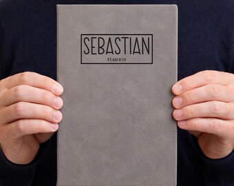 May include: Gray journal with the name "Sebastian" and the last name "Harris" printed in black. The journal is held by a person wearing a navy blue sweater.