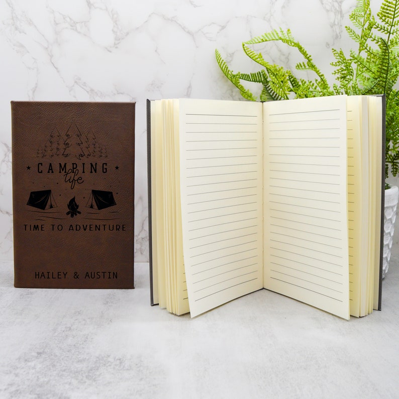 May include: Brown leather-bound journal with a black and white camping themed design. The front cover reads "Camping Life" with a campfire and two tents. The text "Time to Adventure" is below the design. The names "Hailey & Austin" are printed at the bottom of the cover.
