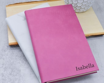 May include: A pink journal with the name "Isabella" printed in black. A white marble-patterned journal is visible beneath the pink journal. The journals are resting on a wooden board.
