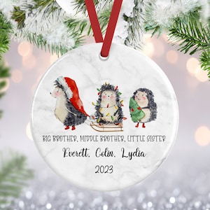 May include: A white marble Christmas ornament with three hedgehogs. The hedgehogs are wearing Santa hats and carrying a Christmas tree and a sled with lights. The text on the ornament reads "BIG BROTHER, MIDDLE BROTHER, LITTLE SISTER Everett, Colin, Lydia 2023".