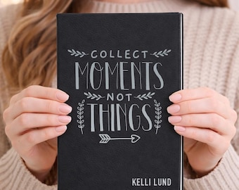 May include: Black journal with the words "COLLECT MOMENTS NOT THINGS" in white script. The journal has a textured cover and the name "KELLI LUND" at the bottom. The journal is held by a person wearing a beige sweater.