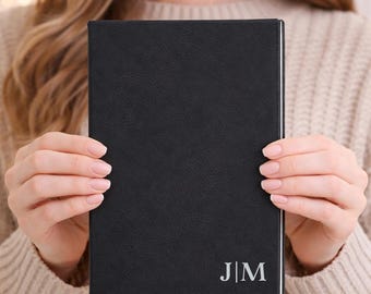 May include: A black journal with a textured cover is held by a person. The journal has the initials "J|M" in silver on the lower right corner. The journal's pages are white, and the cover appears to be made of leather.
