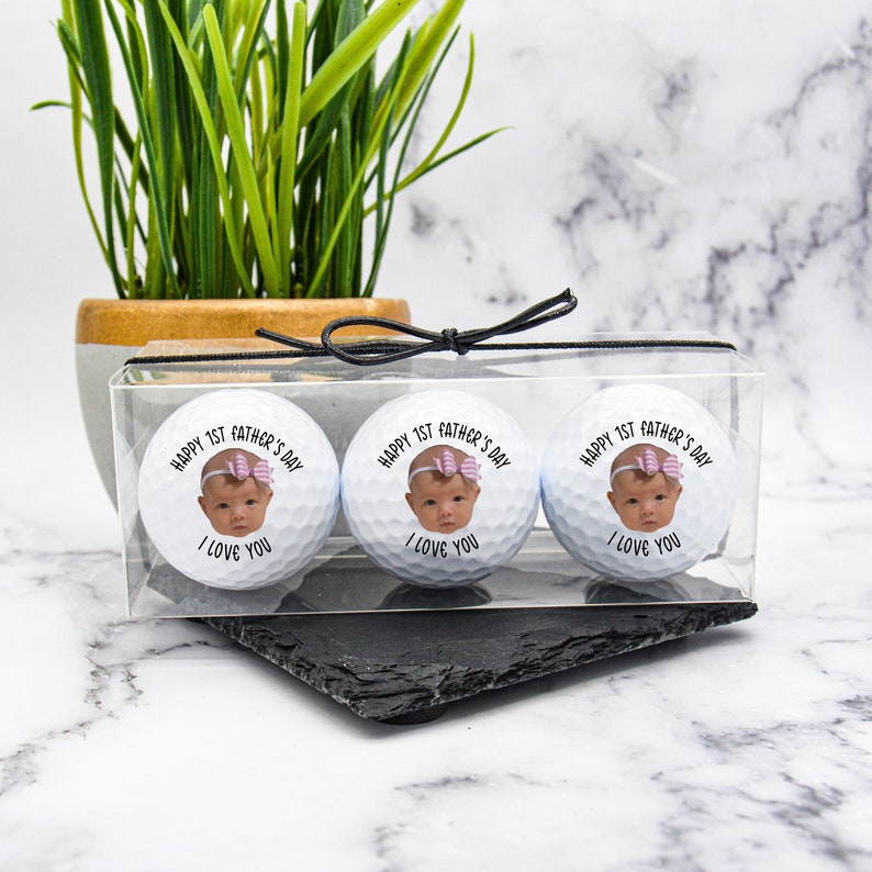 Pu&ograve; includere: Tre palline da golf bianche con una foto personalizzata di una bambina e il testo "Happy 1st Father's Day I Love You" su ogni pallina. Le palline da golf sono in una scatola di plastica trasparente con un nastro nero.