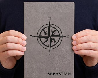 May include: Gray journal with a compass rose design and the name "SEBASTIAN" at the bottom. The compass rose is black and features the cardinal directions. The journal is held by a person wearing a dark blue sweater.