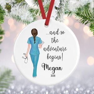 May include: A round white ceramic ornament hanging from a red ribbon. It features an illustration of a person with a braided brown hairstyle wearing light blue scrubs, holding a stethoscope and a clipboard, with the text ...and so the adventure begins Megan 2026.