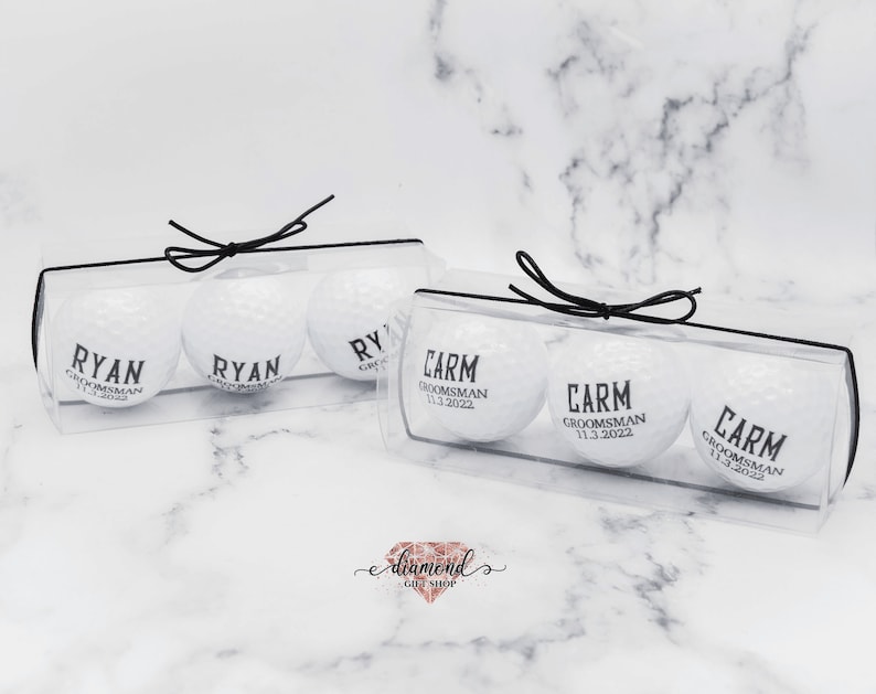 May include: Two clear plastic boxes with black ribbon bows, each containing three white golf balls with black text. The text on the golf balls reads "Ryan,  Groomman" and "Carm, Groomman 11.3.2022".