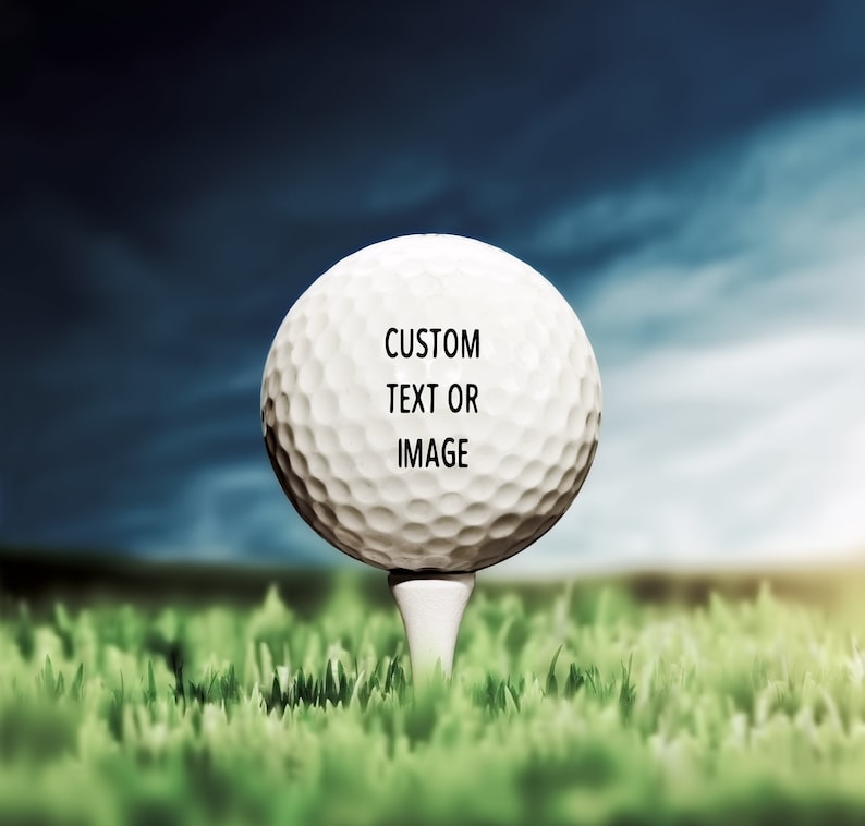 Pode incluir: Uma bola de golfe branca com o texto "CUSTOM TEXT OR IMAGE" nela, sentada em um tee verde em um campo gramado.