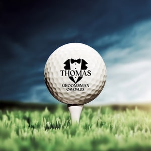 K&ouml;nnte beinhalten: Ein wei&szlig;er Golfball mit einer schwarzen Smoking-Grafik und dem Text "THOMAS GROOMSMAN 09.09.23" darauf gedruckt.