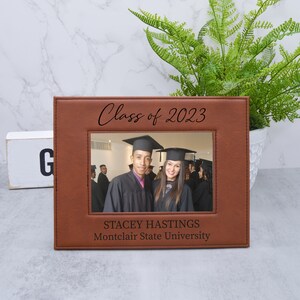 May include: A brown leather picture frame with a photo of two graduates in caps and gowns. The frame has the text "Class of 2023" at the top and "STACEY HASTINGS Montclair State University" at the bottom.