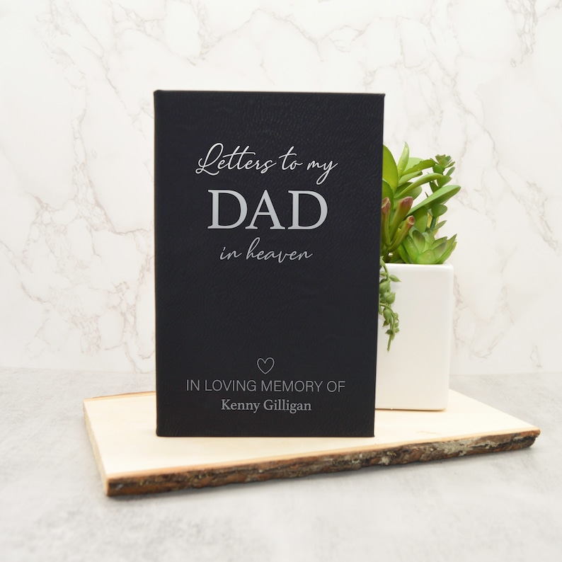 May include: A black journal with the text "Letters to my DAD in heaven" in silver lettering. Below the text is a heart symbol and the inscription "IN LOVING MEMORY OF Kenny Gilligan". The journal is displayed on a wooden slab.