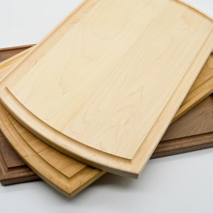 May include: Three wooden cutting boards, one brown and two light wood, with rounded edges and a decorative groove around the perimeter. The boards are stacked on top of each other.