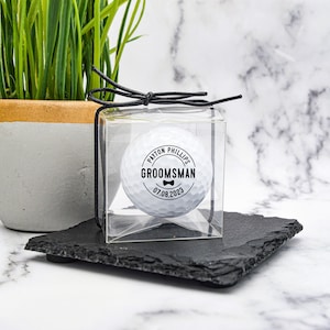 May include: A white golf ball with a black circle sticker that says "PAYTON PHILLIPS GROOMSMAN 07.08.2023" with a bow tie icon. The golf ball is in a clear plastic box with a black ribbon.