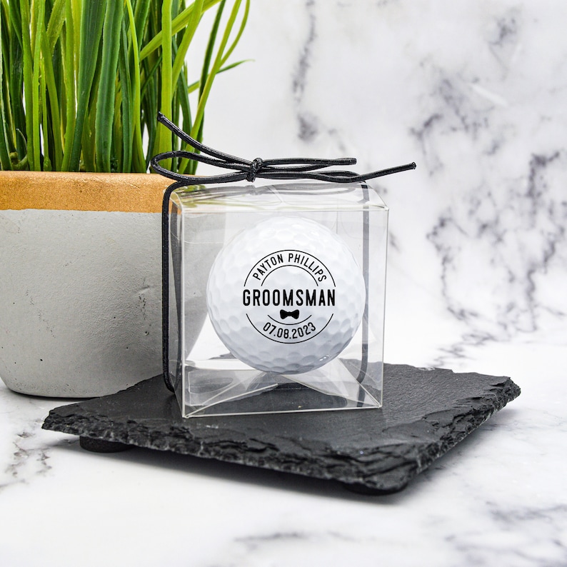 May include: A white golf ball with a black circle sticker that says "PAYTON PHILLIPS GROOMSMAN 07.08.2023" with a bow tie icon. The golf ball is in a clear plastic box with a black ribbon.