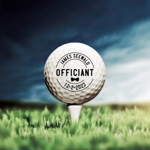 May include: A white golf ball with a black circle on it that says "James Seewald Officiant 12-2-2023" with a small bow tie graphic.