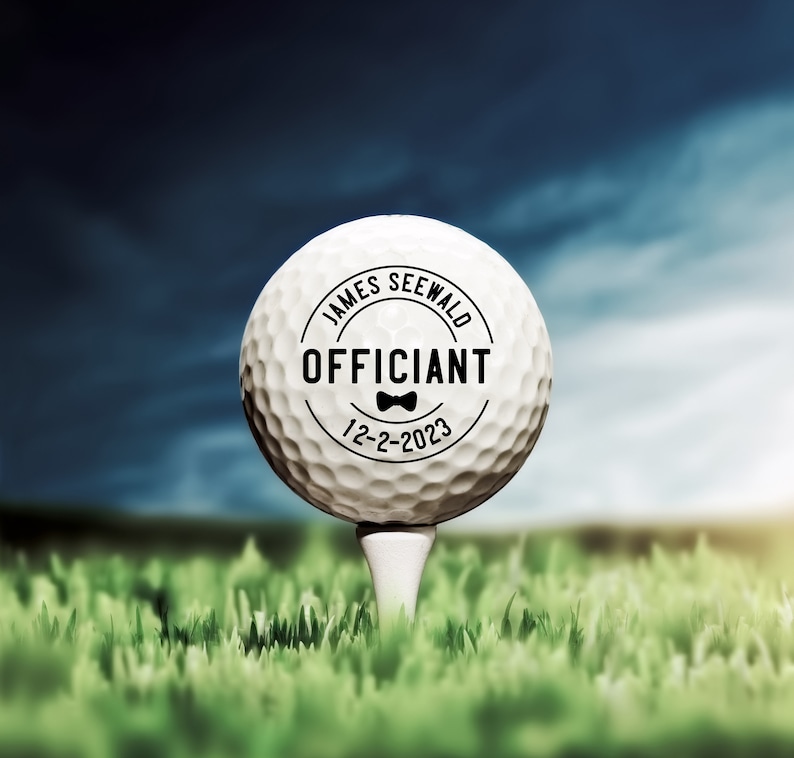 May include: A white golf ball with a black circle on it that says "James Seewald Officiant 12-2-2023" with a small bow tie graphic.
