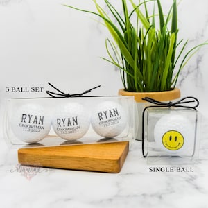 May include: Three white golf balls with black text in clear plastic packaging. The text on the golf balls reads "Ryan,  Groomman, 11.3.2022". A single white golf ball with a yellow smiley face in clear plastic packaging. The text "3 Ball Set" and "Single Ball" are printed below the packaging.