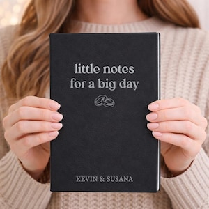 May include: Black journal with the text "little notes for a big day" and two wedding rings. The journal also has the names "KEVIN & SUSANA" printed at the bottom. The journal is held by a person.