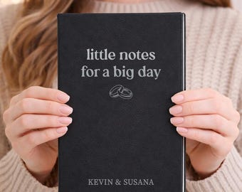 May include: Black journal with the text "little notes for a big day" and two wedding rings. The journal also has the names "KEVIN & SUSANA" printed at the bottom. The journal is held by a person.