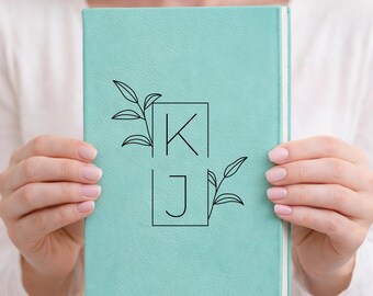 May include: A teal journal with a textured cover. The cover features the initials "K" and "J" within outlined squares, accompanied by leaf designs. The journal is held by a person.