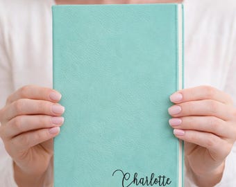 May include: A teal-colored journal with the name "Charlotte" in elegant script. The journal has a textured cover and a white spine. The image shows the journal being held by a person.
