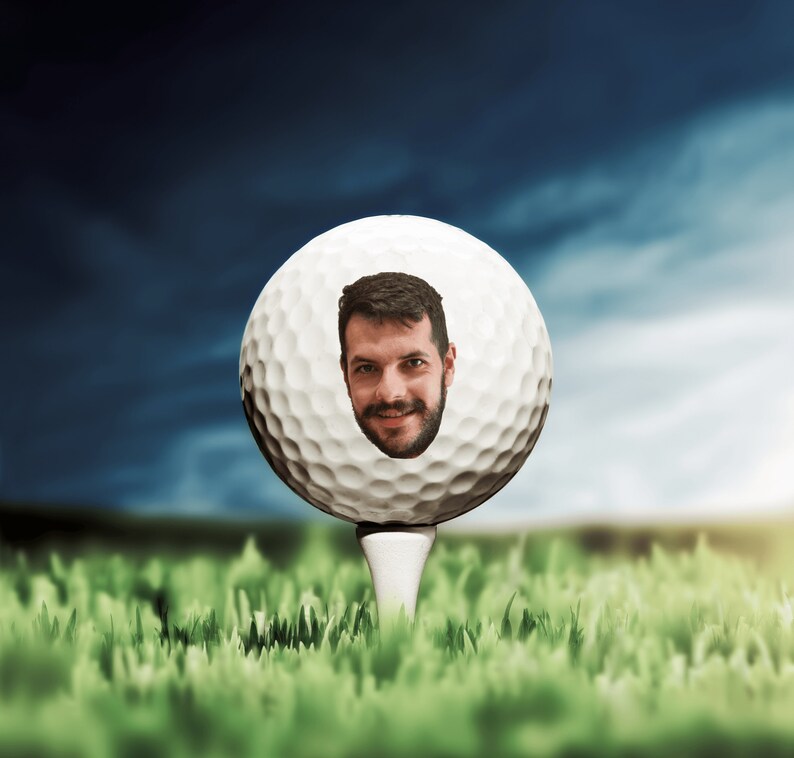 May include: A white golf ball with a man's face in the center, sitting on a tee in a green grassy field.