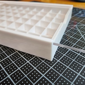 Op de afbeelding: Een witte plastic opbergdoos met een doorzichtig plastic deksel. De doos heeft een raster van vakken om kleine items te organiseren.